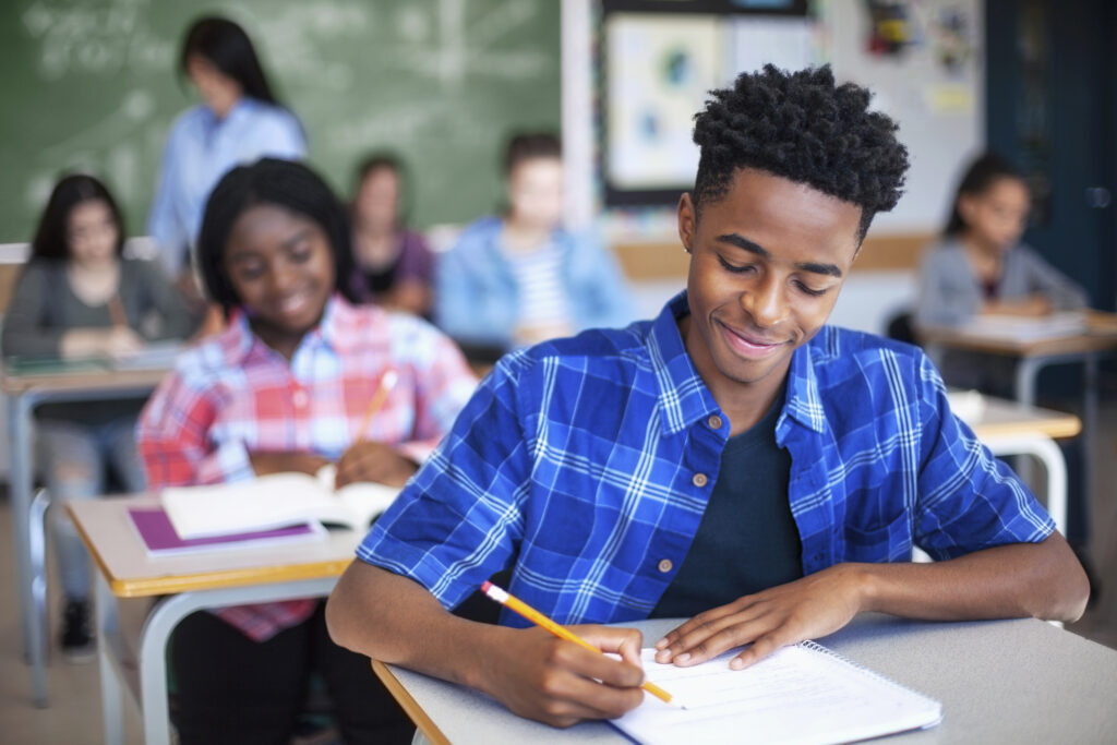 Photo of smiling male student writing in classroom. He is sitting at a desk.