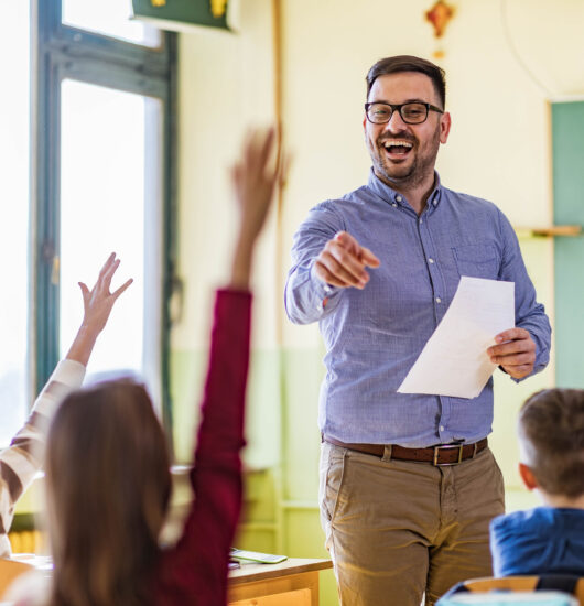 Happy teacher pointing at his elementary-aged students who raised their hands to answer a question in a class.