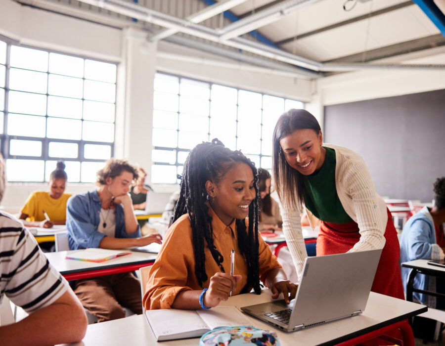 Photo of smiling young woman teacher helping a student working on a laptop during a lesson in a classroom