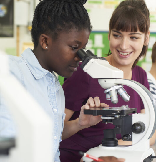 Photo of teacher looking on as a student peers through the lens of a microscope.