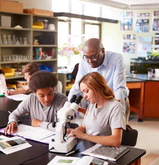 High School Students With Tutor Using Microscope In Biology Class