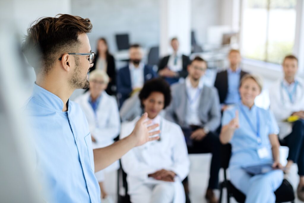 Man wearing a blue button up and glasses talking to a room full of other well-dressed people.